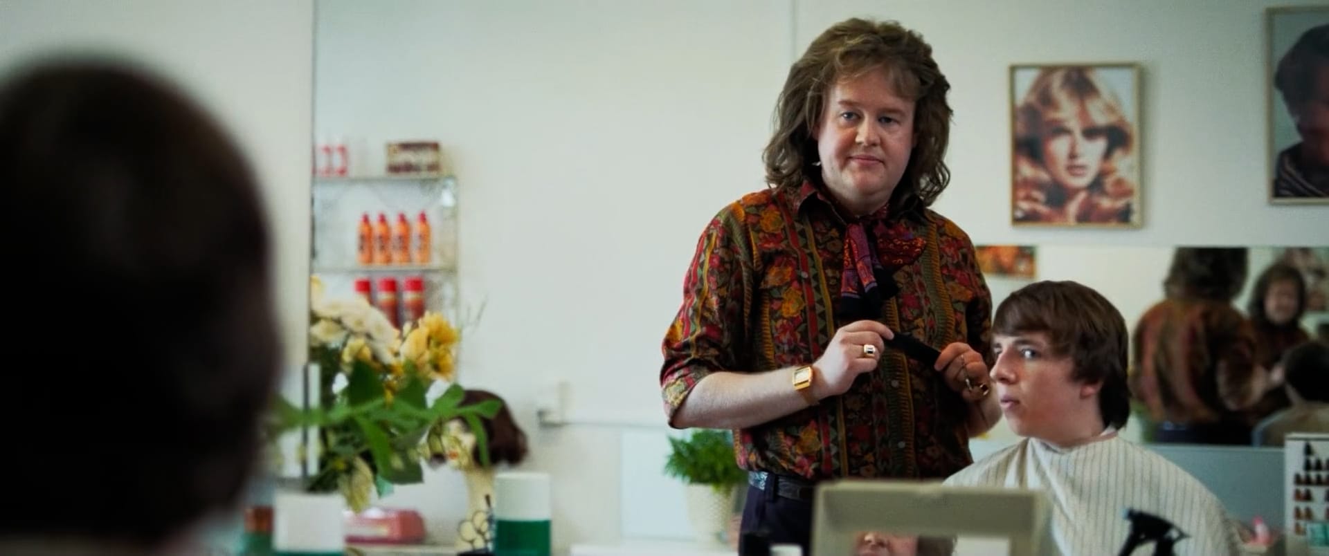 A male 1970s hairdresser and a teenage boy looking in the mirror. The hairdresser has long voluminous hair and is wearing a colourful shirt and tied scarf and an ostentatious ring and digital watch. He is holding a comb. The boy is mid-haircut. He has longish hair and is unsure of the haircut. In the background are hair products, an image of a lady's hairstyle, flowers, and a reflection of a mirror showing a reflection of the reflection.